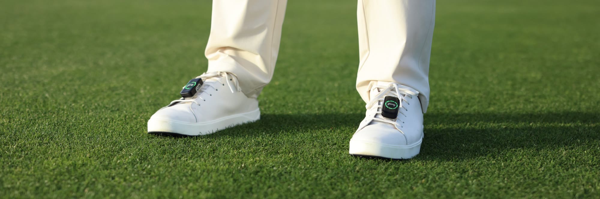 A golfer standing on a fairway in white shoes, with a Laced sensor clipped to the laces of each shoe and its ring lit green.